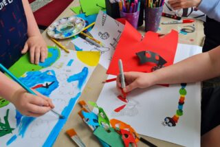Children's hands engaged in creative art, painting and colouring with vibrant papers and markers on a tabletop covered in artwork.