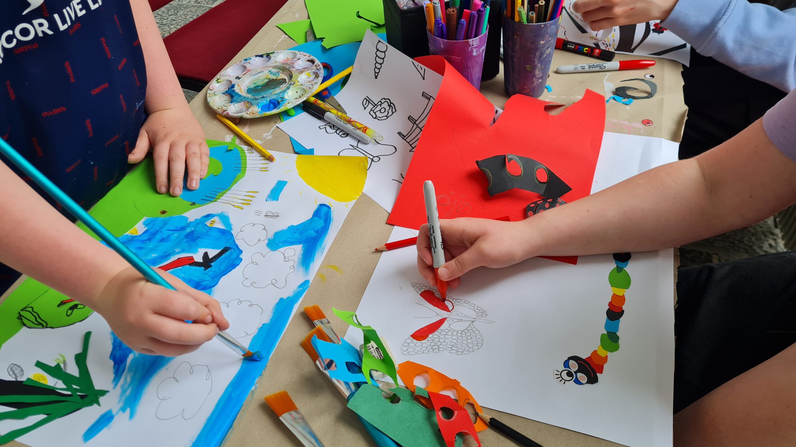Children's hands engaged in creative art, painting and colouring with vibrant papers and markers on a tabletop covered in artwork.