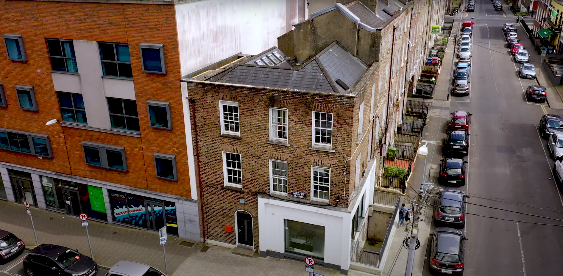 Aerial view of a brick building between modern structures, with parked cars lining the street and pedestrians walking nearby.