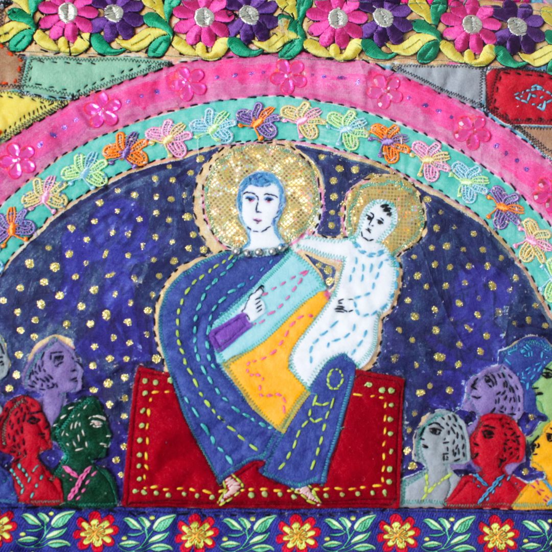 A vibrant textile artwork depicting a seated figure with a child, surrounded by intricate floral patterns and colourful faces in a collage.