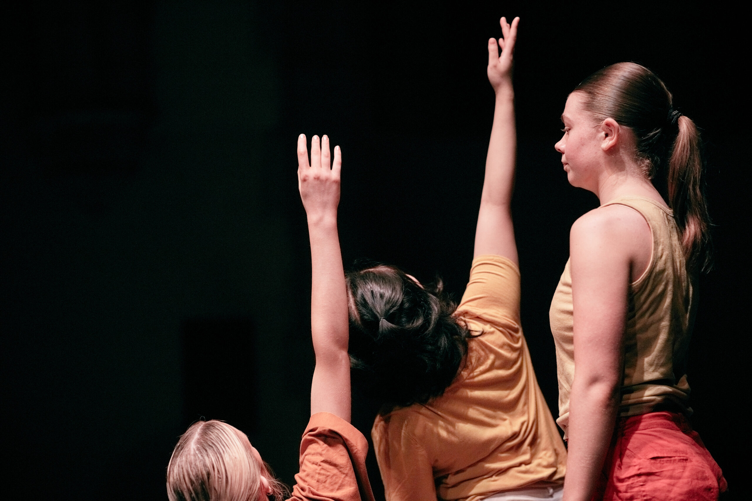 Three dancers in warm-coloured shirts and pants perform under spotlight, arms raised upwards, conveying focus and unity against a dark background.