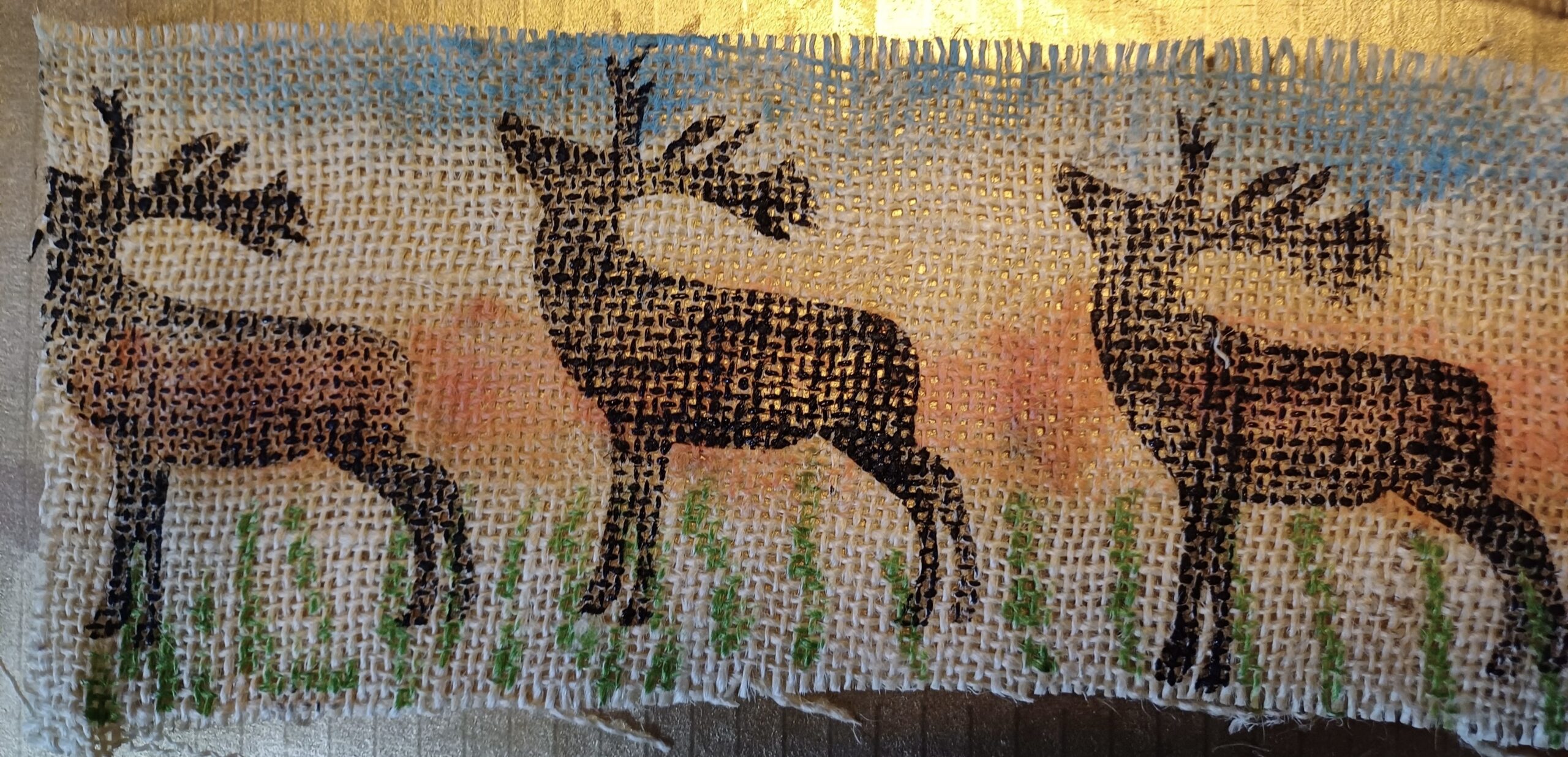 Three black deer silhouettes on woven burlap, with a gradient sky backdrop in blue and orange hues. The foreground features green grass accents.