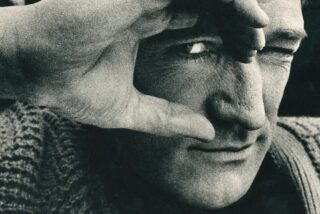 Black and white close-up of Richard Harris's face, one eye visible through fingers framing it. Textured sweater in foreground, creating a contemplative mood.