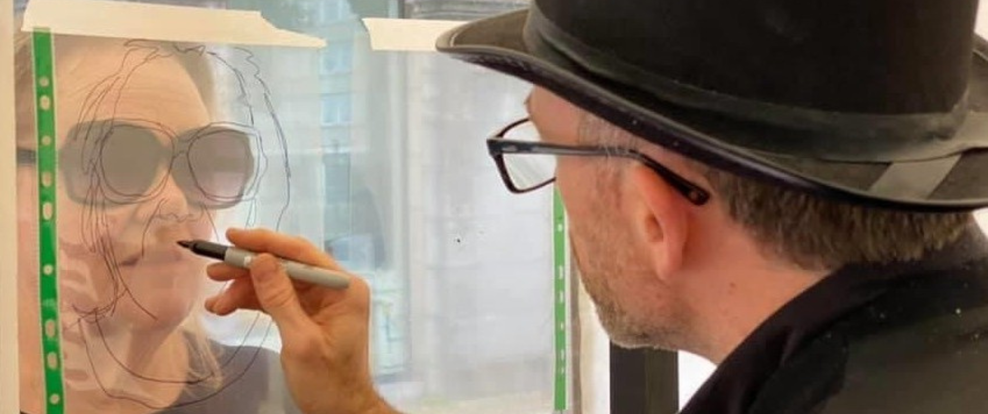 A close up shot of a male artist wearing a hat drawing a woman wearing sunglasses.