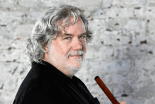 A man with grey hair is holding a flute, poised to play, with a focused expression on his face.