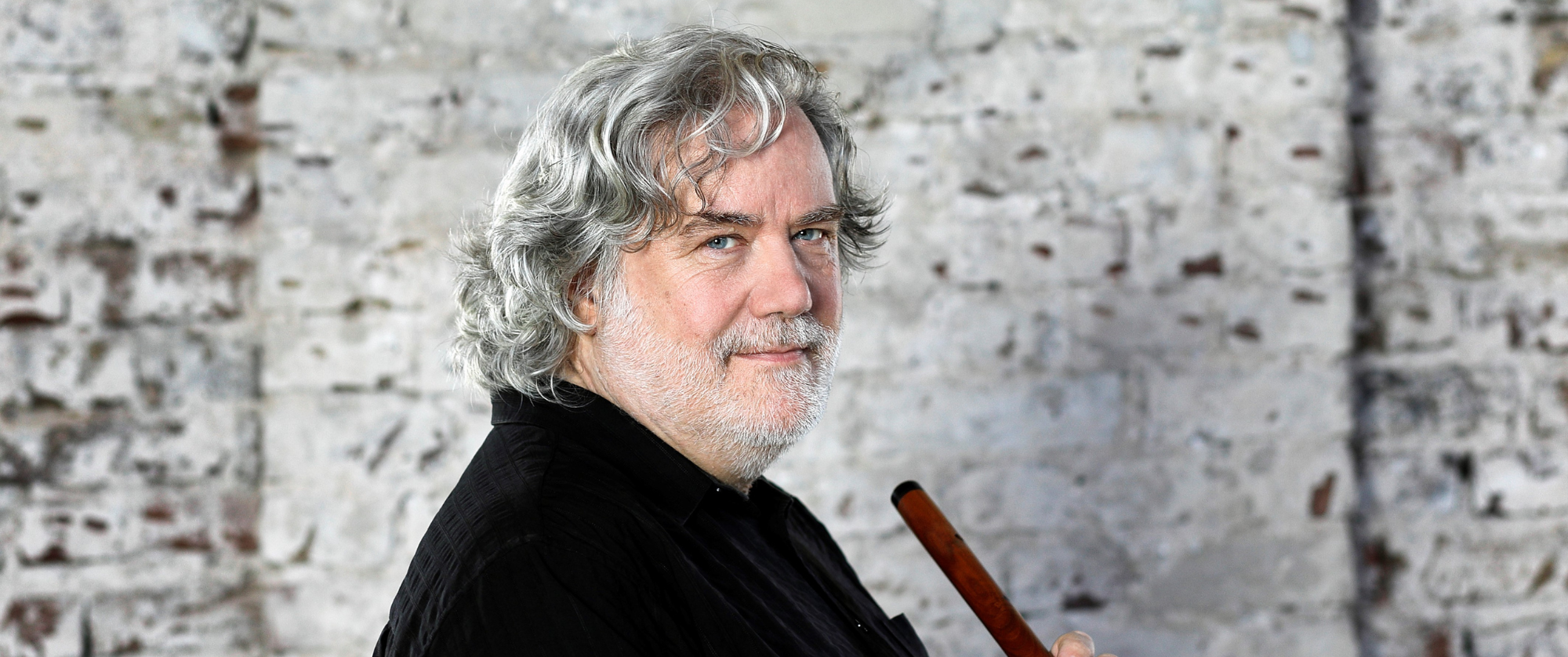 A man with grey hair is holding a flute, poised to play, with a focused expression on his face.