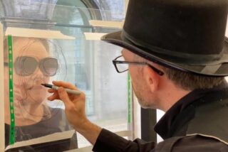 A male artist wearing a hat drawing a woman wearing sunglasses.