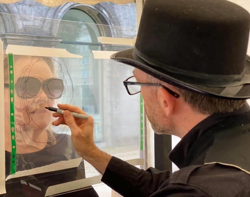 A male artist wearing a hat drawing a woman wearing sunglasses.